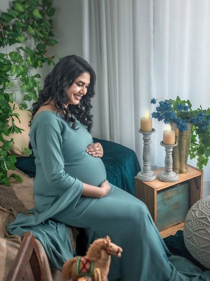 A smiling, happy portrait of the mother-to-be cradling her bump. The natural light from the window and the soft green tones of the decor create a fresh and joyful mood.
