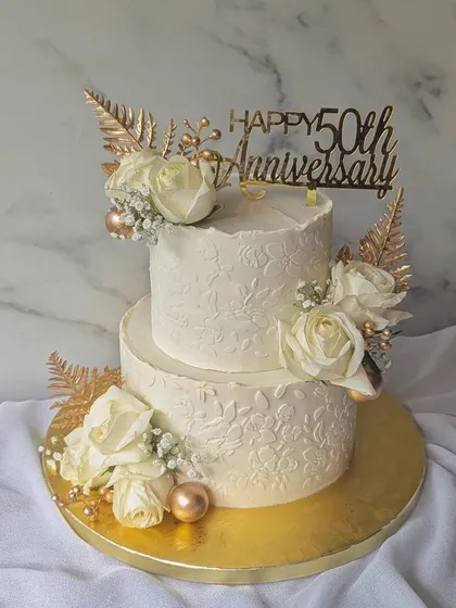 Celebrating a golden anniversary with this elegant two-tier cake. The design features a subtle floral stencil, white roses, and striking gold-dusted ferns for a touch of glamour.