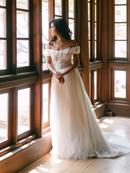 A bride in her beautiful off-the-shoulder gown, looking out a large window. The natural light illuminates her thoughtful expression, creating a classic and romantic bridal portrait.