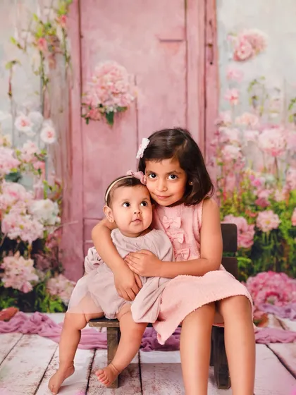 A classic portrait of an older sister holding her baby sister on a small chair. The floral background adds a touch of magic to this sweet moment.