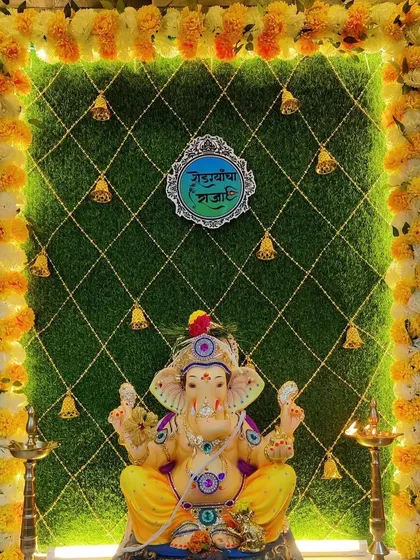 A creative Ganpati backdrop using a green turf wall with a crisscross pattern and hanging bells. The frame of marigold flowers adds a traditional touch to this unique design.