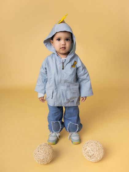A full-length portrait of a toddler standing confidently in a cute blue hooded jacket against a solid yellow background. This minimalist setup makes his adorable outfit and expression pop.