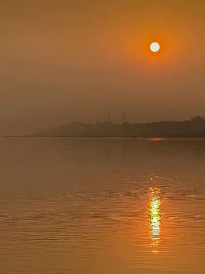A hazy sunrise over a large body of water, with the sun's reflection creating a bright column of light. The mist gives the scene a soft, dreamy quality.