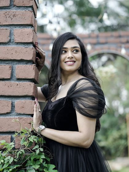 A lovely solo portrait of the bride-to-be in a black tulle gown, smiling by a brick pillar in a garden setting.