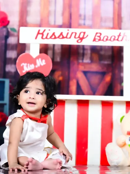 A baby girl sitting on the floor of the "Kissing Booth" setup, looking up with a curious expression.