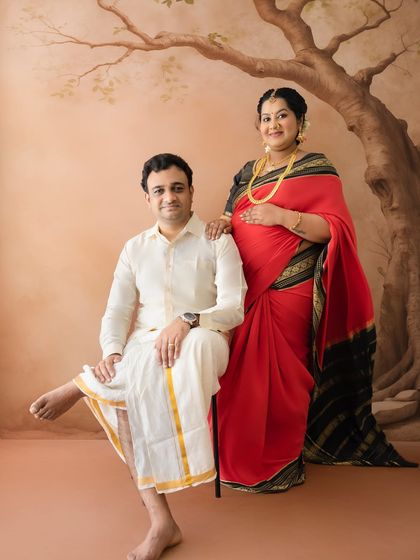 A poised and elegant couple's portrait. The mom-to-be stands gracefully while her partner is seated, both in traditional attire, creating a balanced and regal composition.