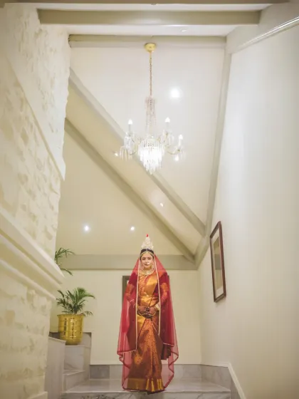 A full-length portrait of a Bengali bride standing at the top of a staircase. The grand chandelier and minimalist decor create a regal and elegant setting.