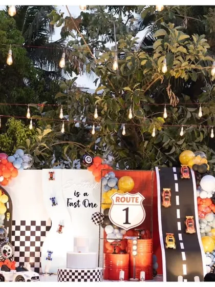 An action-packed "Fast One" first birthday party with an outdoor setup. The decor features a race track prop with toy cars, a Route 1 sign, and checkered flag details.