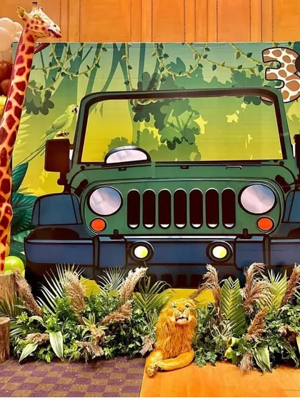 A full view of the safari jeep photo booth, surrounded by jungle foliage and a tall giraffe prop. The balloon garland adds a touch of festive color to the wild green and brown palette.