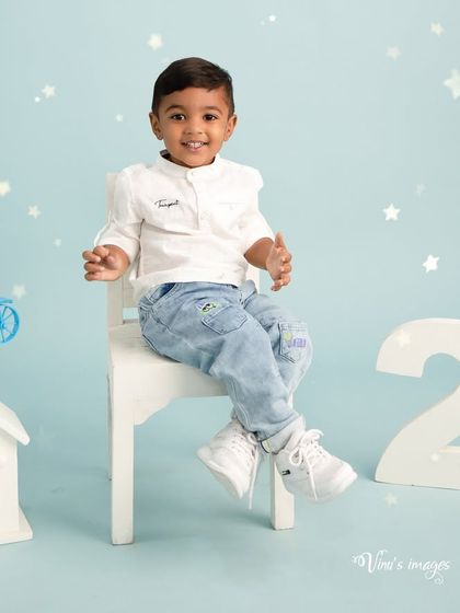 A cool and casual portrait for a second birthday. The birthday boy sits confidently on a small white chair, smiling for the camera in this clean and simple studio setup.