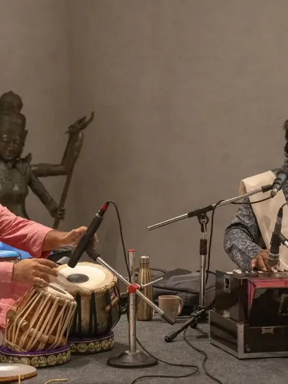 Providing tabla accompaniment for a harmonium player and vocalist in a chamber music setting. The statue of Nataraja in the background adds to the artistic ambiance.