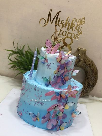 A pretty two-tier butterfly-themed cake for an 8th birthday. This 3kg chocolate mousse cake is covered in blue whipped cream and a cascade of colorful, edible butterflies, complete with a custom name topper.