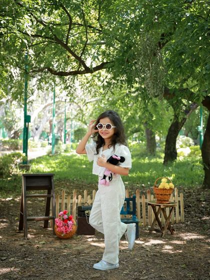 A full-length shot of a stylish girl posing in a park. The natural, dappled sunlight filtering through the trees creates a beautiful lighting effect for outdoor portraits.