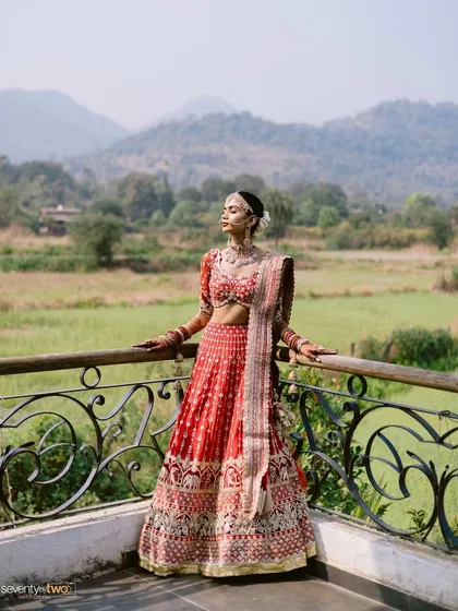 A bride stands on a balcony overlooking a vast green landscape, her red lehenga a vibrant splash of color against the serene natural backdrop.