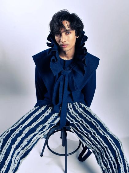 A seated portrait of a male model in a dramatic, high-collar shirt. The pose is confident and styled for a high-fashion context.