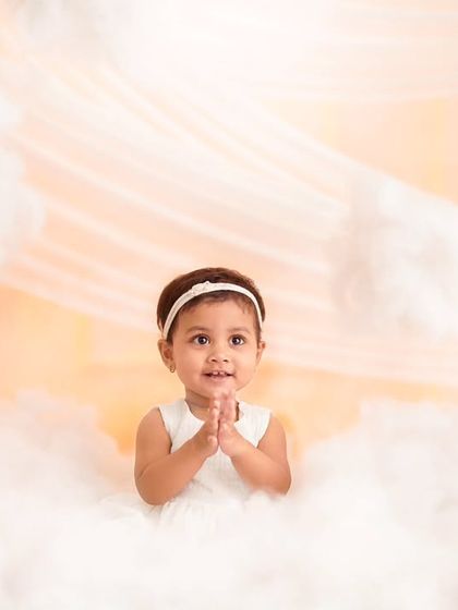 A sweet toddler in a white dress surrounded by soft, cloud-like props, looking up with a gentle expression.