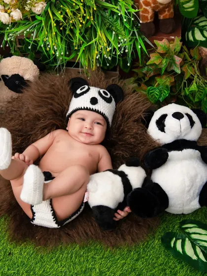 A smiling baby is dressed in an adorable hand-knitted panda costume, lying next to a plush panda toy. This playful animal theme is a fun way to capture your baby's delightful personality.