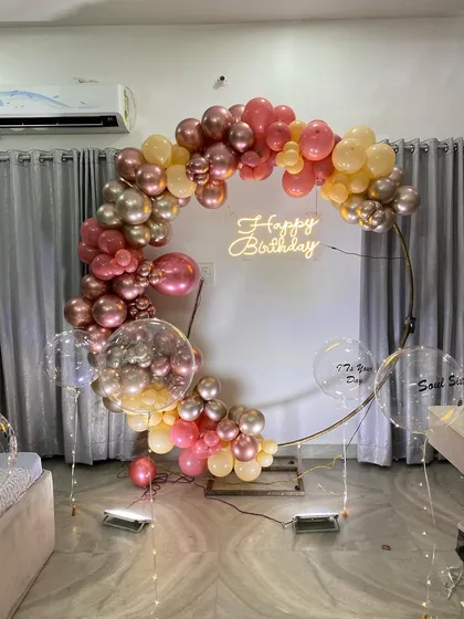 This is a duplicate of ID 17, 49, 92, 153, 186. An elegant balloon ring decoration for an intimate home celebration. This setup features a half-arch of chrome and pastel balloons on a golden ring, with a 'Happy Birthday' neon sign.