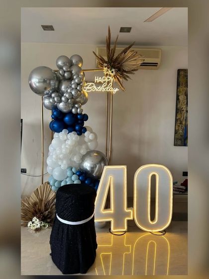Here's to 40! This stylish setup features a modern gold frame with a blue, silver, and white balloon cascade. A "Happy Birthday" neon sign, large "40" LED numbers, and dried floral arrangements complete this contemporary and masculine design.