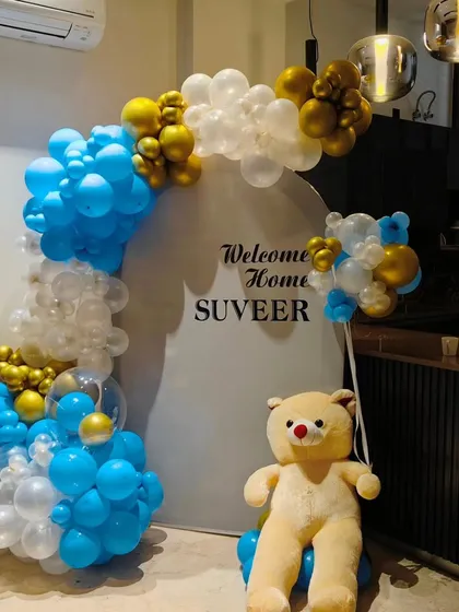 For a modern and clean 'Welcome Home' celebration, I created this setup for baby Suveer. It features a stylish blue, white, and gold balloon arch against a simple white circular backdrop, complemented by a cute teddy bear.