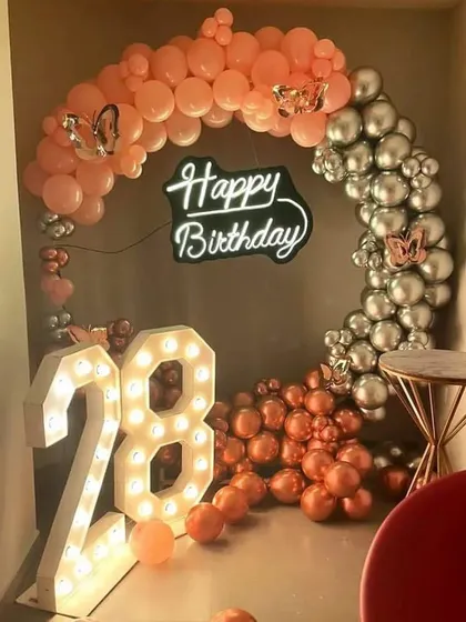 A 28th birthday celebration with a split-color balloon arch in peach and silver, a 'Happy Birthday' neon sign, and a '28' marquee light.
