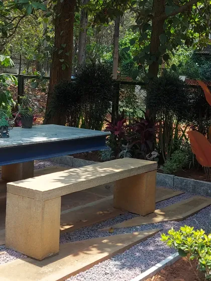 A secluded outdoor dining area with a concrete table and stone bench. The space is nestled among trees and features a gravel ground cover, creating a rustic and natural feel.