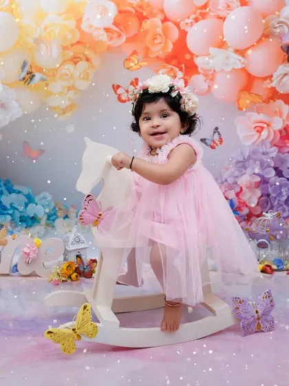 A smiling girl on a rocking horse, surrounded by pastel balloons and butterflies for her magical first birthday.