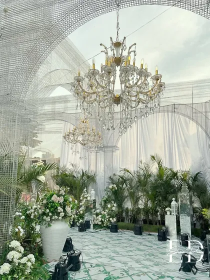 A breathtaking view of a wedding venue transformed with custom-built wire mesh arches and magnificent crystal chandeliers. The space is filled with potted palms and white floral arrangements, creating a sustainable botanical garden feel.