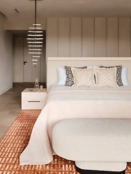 A serene bedroom composition featuring a statement pendant light that acts as a sculptural element. The design combines soft textures, a warm, earthy rug, and clean lines to achieve a state of cozy minimalism.