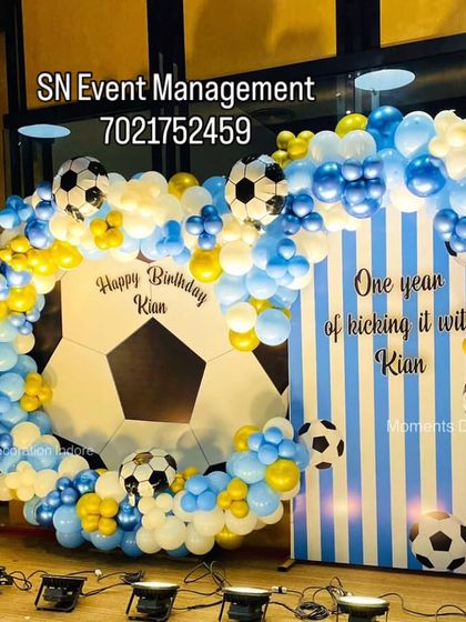 A unique football-themed birthday decoration for a first birthday. The setup includes a goal-like balloon arch with football balloons and a custom backdrop celebrating "One year of kicking it with Kian".