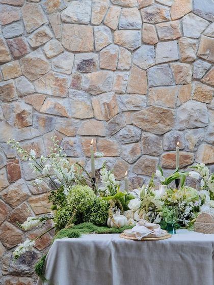 A wider view of the Earth Table, showcasing the gentle flicker of candles among the green and white floral arrangements. The natural stone wall provides a perfect rustic backdrop.