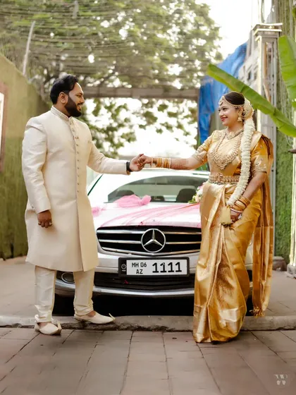 A full-length shot of the couple with their decorated wedding car, showcasing their elegant South Indian wedding attire.