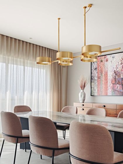 The dining area in the Maximum City Duplex features a marble-topped table and chairs upholstered in a dusty rose fabric. The brass chandelier above adds a final touch of elegance.