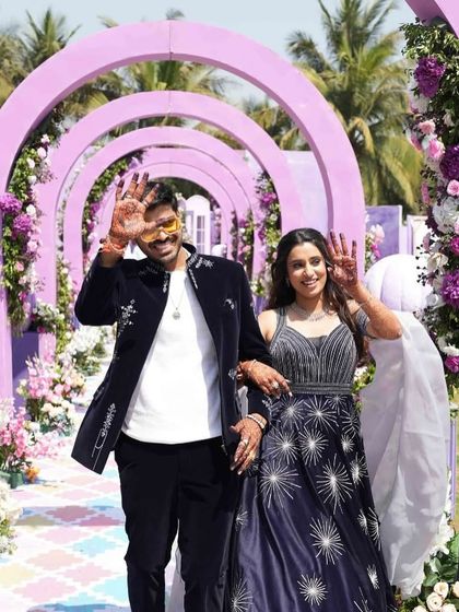The happy couple, framed by the pastel arches of the carnival entrance, showing their henna-adorned hands.