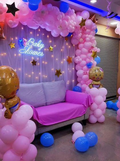 A full-room baby shower decoration for a home party. The setup includes a 'Baby Shower' neon sign against a curtain with fairy lights, and extensive balloon decorations in pink and blue, including character foil balloons.