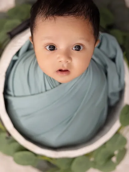 A striking close-up shot from above, this newborn's wide eyes are the center of attention. The simple teal swaddle and leafy green accents create a clean and engaging portrait.