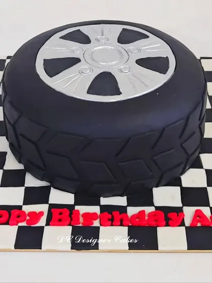 This car tyre cake was designed for a car lover's birthday. The realistic tread and silver rim are crafted from fondant, sitting on a checkered flag board to complete the racing theme.