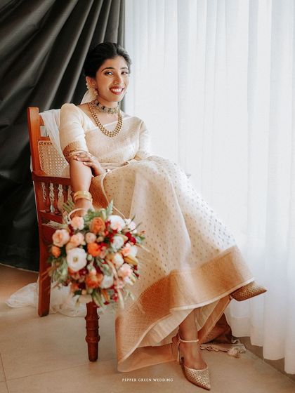 A stunning portrait of the bride seated by a window, her elegant white and gold saree and bouquet creating a picture of grace.
