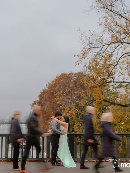 An artistic shot using slow shutter speed to blur the motion of people walking past the couple on a Parisian bridge. The couple remains still in their embrace, a calm center in the midst of the city's hustle.
