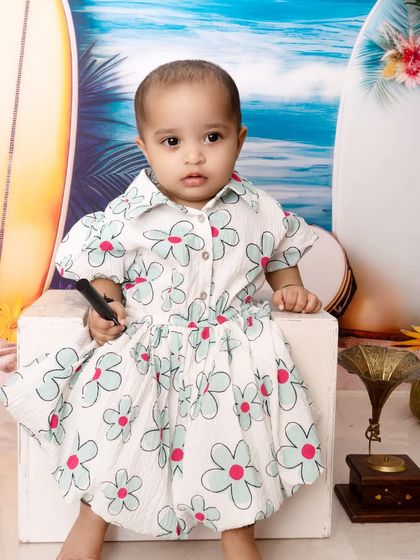 A close-up portrait from a beach-themed session. The baby is dressed in a floral outfit, sitting against a backdrop of the ocean and surfboards for a fun, summery vibe.