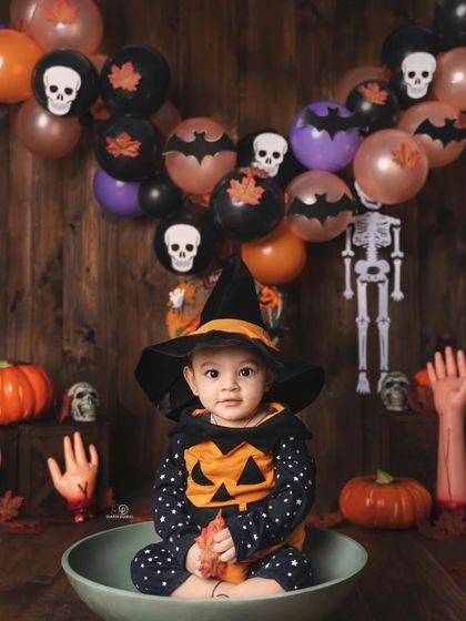 A spooky and fun Halloween-themed photoshoot. This baby, dressed in a witch hat and pumpkin outfit, crawls out of a bowl amidst pumpkins, skeletons, and other festive decorations.