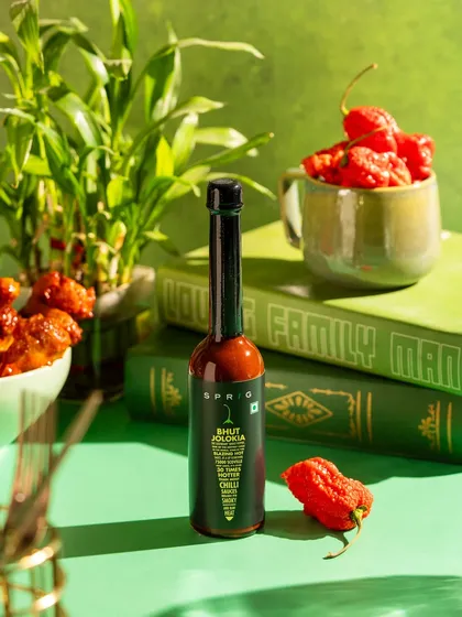 A product shot for Sprig's Bhut Jolokia hot sauce. The vibrant green background and fresh ghost peppers communicate the product's key ingredient and heat.