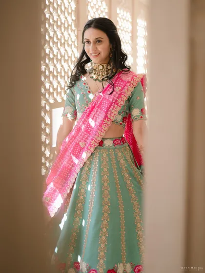 A glimpse of sunshine. Peeking through a traditional jali window, this portrait captures the bride's happy smile. Her look is styled with a vibrant pink bandhani dupatta, adding a pop of color to her pastel lehenga.