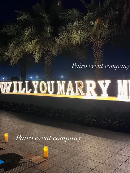 A close-up of the "WILL YOU MARRY ME" marquee letters, showing their bright illumination against the night sky and palm trees, perfect for a destination proposal feel.