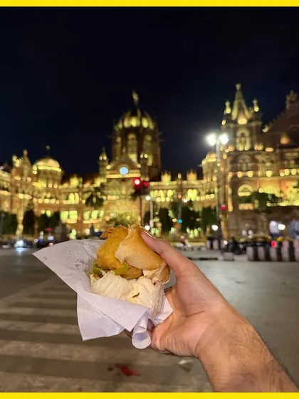 A vada pav held up against the illuminated CSMT station at night, a delicious contrast of street food and grand architecture.