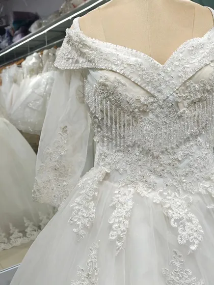 A stunning off-the-shoulder ball gown with intricate beadwork and fringe details on the bodice. The long, sheer sleeves are embroidered to match, creating a cohesive and luxurious look.