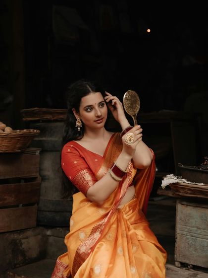 A beautiful portrait of a woman in a Banarasi saree, getting ready in a traditional setting, using a brass mirror to check her appearance.