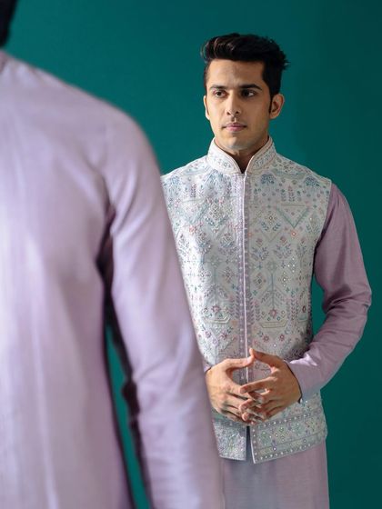 A model is reflected, wearing a stunning raw silk Jawahar jacket with intricate mirror and threadwork. This piece, in a beautiful lilac and silver palette, is ideal for a Mehendi or Pooja function.