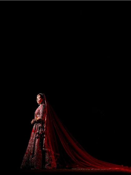 A breathtaking full-length shot against a black background. The bride's red lehenga with its long trail creates a silhouette of pure drama and elegance.