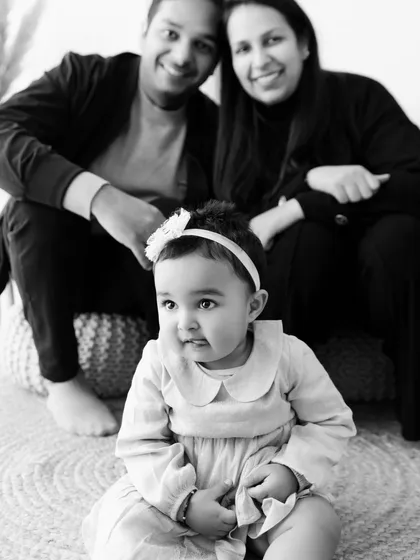 A black and white photo of a baby in the foreground with her parents smiling behind her. This composition creates a lovely, layered family portrait.